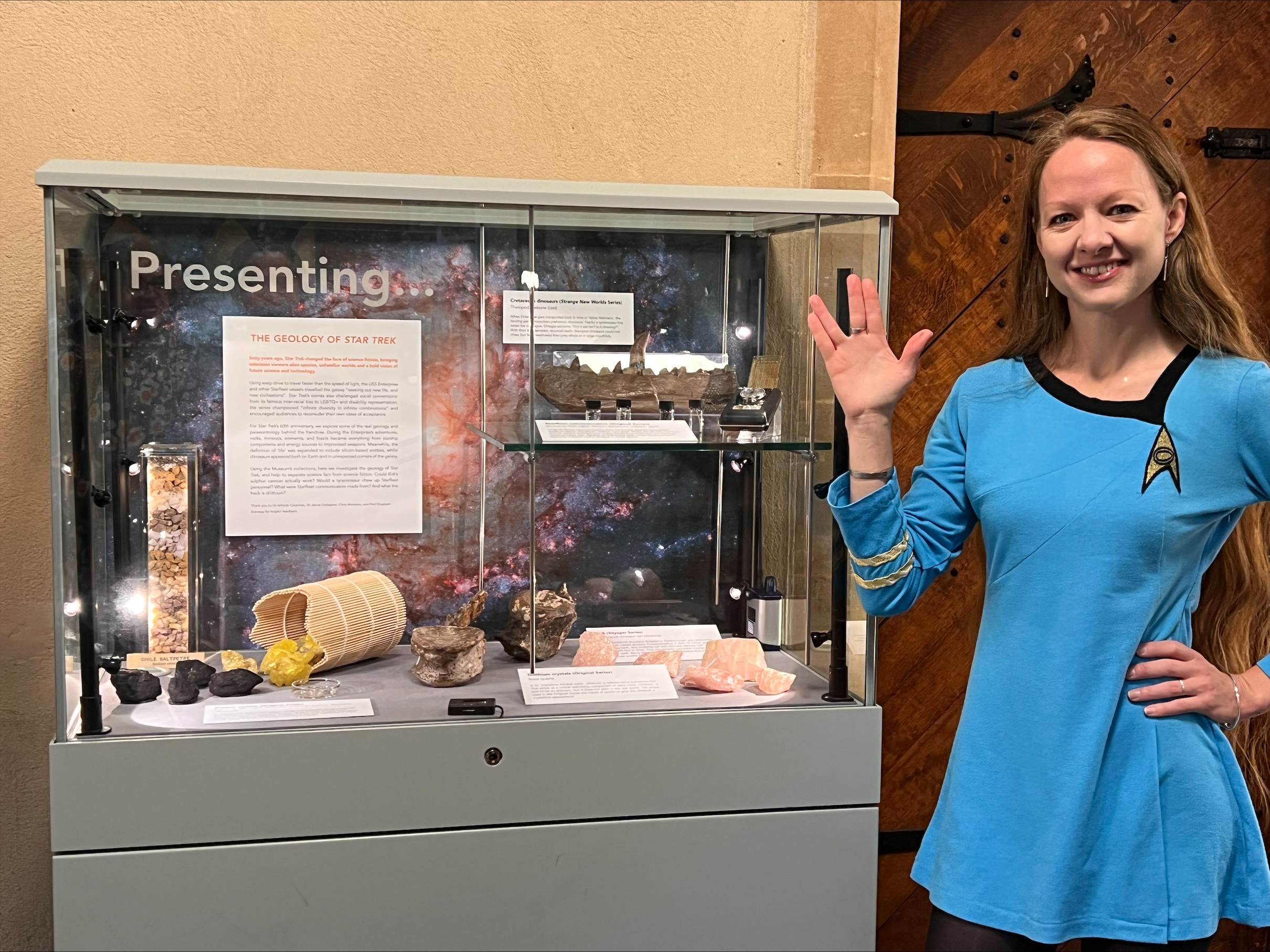 woman wearing a blue Starfleet uniform cosplay from Star Trek The Original series, doing a Vulcan salute, standing next to an exhibit case with rock samples and labels discussing the geology of Star Trek woman wearing a blue Starfleet uniform cosplay from Star Trek The Original series, doing a Vulcan salute, standing next to an exhibit case with rock samples and labels discussing the geology of Star Trek